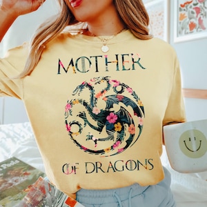 May include: A pale yellow crewneck t-shirt featuring the text MOTHER OF DRAGONS in black lettering. Centered between the words is a circular graphic of a three-headed dragon filled with a dark green tropical leaf and colourful floral pattern.