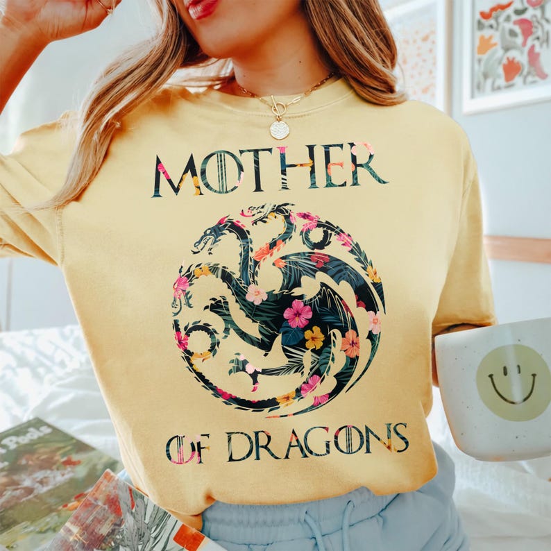 May include: A pale yellow crewneck t-shirt featuring the text MOTHER OF DRAGONS in black lettering. Centered between the words is a circular graphic of a three-headed dragon filled with a dark green tropical leaf and colourful floral pattern.