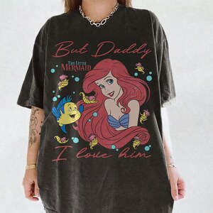 May include: A charcoal gray t-shirt featuring a graphic print of Ariel from The Little Mermaid with red hair, a blue seashell top, and the yellow fish Flounder. The text reads But Daddy I love him in pink script lettering with small seahorses.