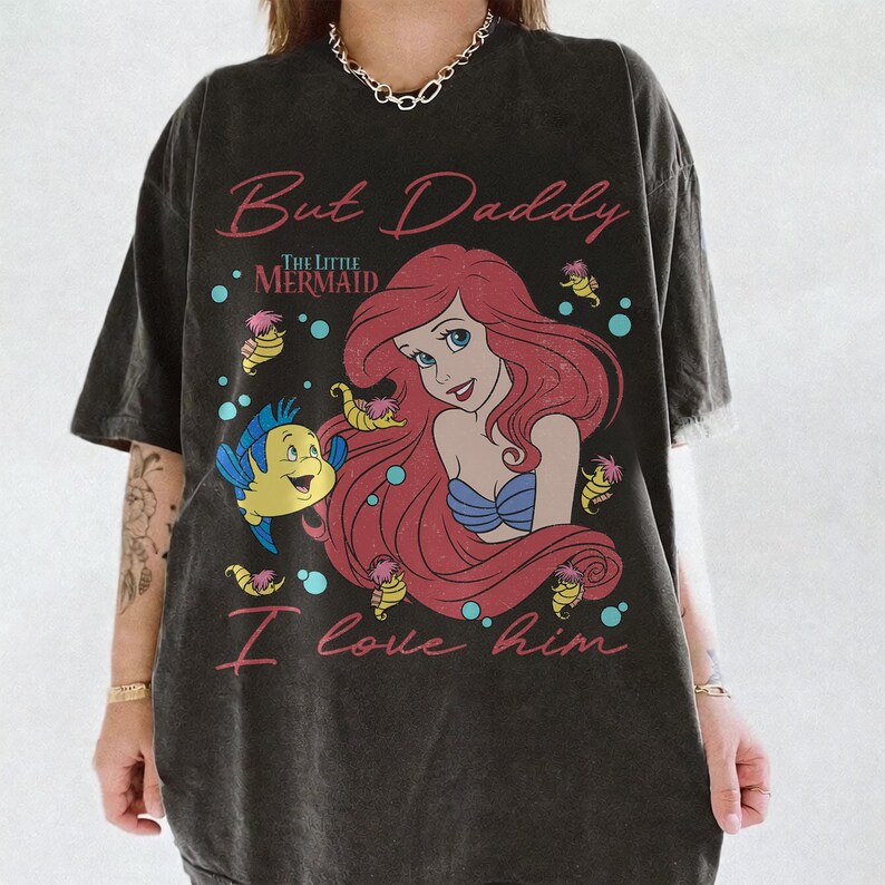 May include: A charcoal gray t-shirt featuring a graphic print of Ariel from The Little Mermaid with red hair, a blue seashell top, and the yellow fish Flounder. The text reads But Daddy I love him in pink script lettering with small seahorses.
