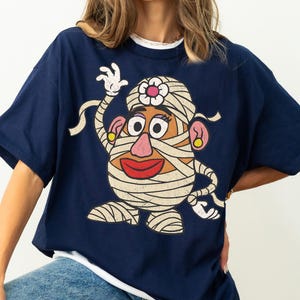 May include: A navy blue short-sleeved t-shirt featuring a large graphic print of a potato character wrapped in white bandages like a mummy. The character has a pink nose, red lips, yellow earrings, and a white flower on its head, with a distressed texture.