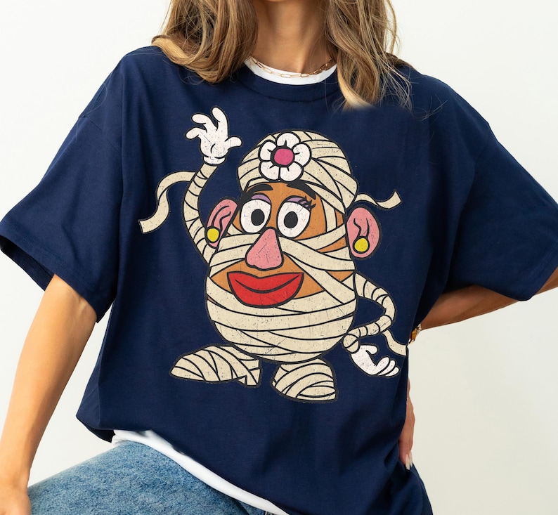 May include: A navy blue short-sleeved t-shirt featuring a large graphic print of a potato character wrapped in white bandages like a mummy. The character has a pink nose, red lips, yellow earrings, and a white flower on its head, with a distressed texture.
