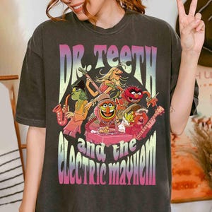 May include: Dark gray t-shirt featuring a colorful graphic of Dr. Teeth and the Electric Mayhem band. The design includes the band's name and images of the Muppet characters playing instruments. The shirt has a vintage, faded look.