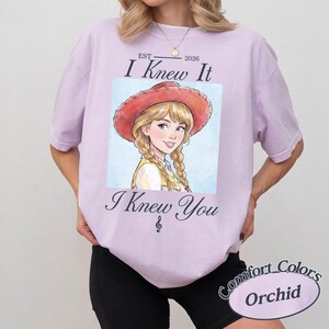 May include: A light purple t-shirt featuring a graphic of a blonde character wearing a red cowboy hat with braided hair. The text reads EST 2026 I Knew It I Knew You with a musical treble clef symbol below. The shirt has short sleeves and a crew neck.