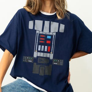 Op de afbeelding: Een marineblauw t-shirt met korte mouwen en een grafische print van een mechanische borstplaat. Het ontwerp bevat grijze panelen, rode en blauwe rechthoekige knoppen en een centrale bedieningsconsole met diverse geometrische vormen.
