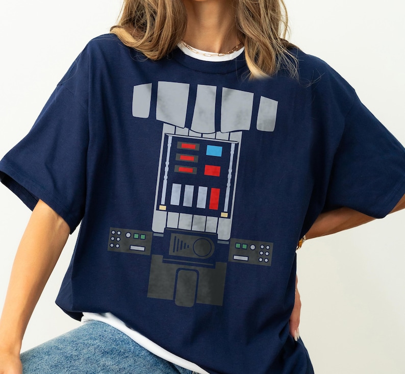 Op de afbeelding: Een marineblauw t-shirt met korte mouwen en een grafische print van een mechanische borstplaat. Het ontwerp bevat grijze panelen, rode en blauwe rechthoekige knoppen en een centrale bedieningsconsole met diverse geometrische vormen.