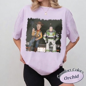 Op de afbeelding: Een lichtpaars t-shirt met korte mouwen en een grafische print van Woody en Buzz Lightyear die op de motorkap van een auto zitten. Woody houdt een sigaret vast en Buzz houdt een groen energiedrankje vast. De nummerplaat toont de tekst BR ER.