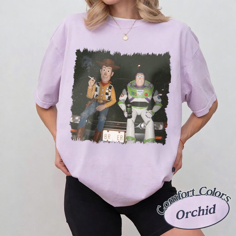 Op de afbeelding: Een lichtpaars t-shirt met korte mouwen en een grafische print van Woody en Buzz Lightyear die op de motorkap van een auto zitten. Woody houdt een sigaret vast en Buzz houdt een groen energiedrankje vast. De nummerplaat toont de tekst BR ER.