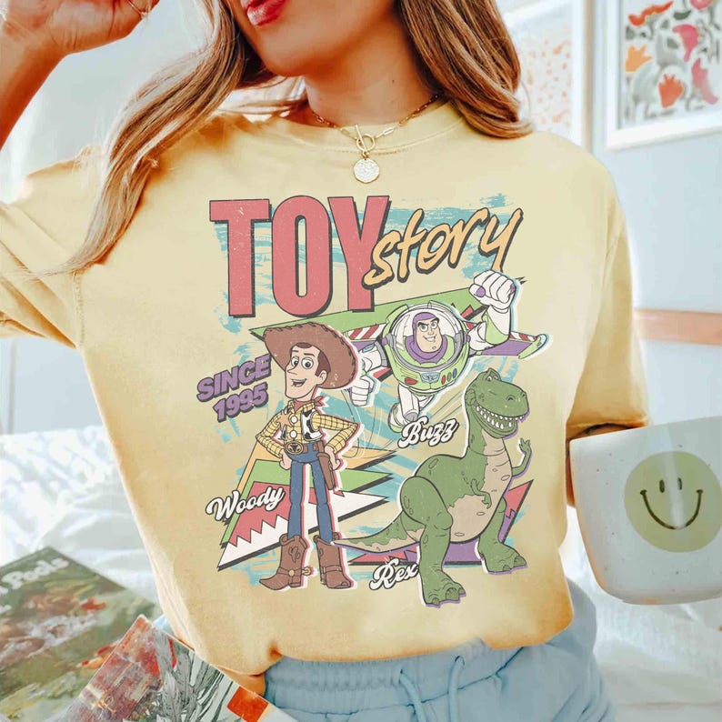 May include: Yellow t-shirt featuring a graphic of Toy Story characters Woody, Buzz Lightyear, and Rex. The design includes the text "TOY STORY" in pink and blue, with "SINCE 1995" below. The shirt has a vintage, retro style.