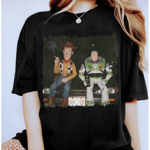 Op de afbeelding: Een zwart t-shirt met korte mouwen en een grafische print van Woody en Buzz Lightyear die op de motorkap van een auto zitten. Woody houdt een sigaret vast en Buzz een Monster-energiedrankje. De nummerplaat toont de tekst BR ER.