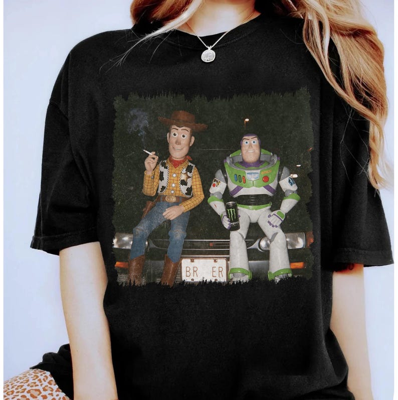 Op de afbeelding: Een zwart t-shirt met korte mouwen en een grafische print van Woody en Buzz Lightyear die op de motorkap van een auto zitten. Woody houdt een sigaret vast en Buzz een Monster-energiedrankje. De nummerplaat toont de tekst BR ER.