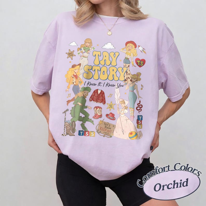 May include: A light purple short-sleeve t-shirt featuring a graphic print with the text TAY STORY I Knew It, I Knew You. The design includes various illustrated figures, a red cardigan, cowboy boots, a hat, a sign reading TAY'S ROOM KEEP OUT, and blocks.