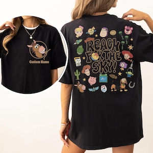 May include: A black t-shirt featuring a front graphic of a horse head inside a rope circle with the text Custom Name. The back displays various cartoon character icons and the text REACH FOR THE SKY! in a large, bold, brown, wavy font.