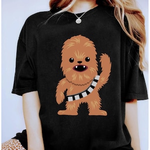 May include: Black t-shirt with a cartoon image of Chewbacca. The brown and tan character has a black and white bandolier and is waving. The shirt is made of a soft material.
