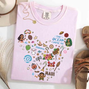 May include: Pale pink t-shirt with a heart-shaped design featuring cartoon characters and the text "MOANA" and "MAUI". The shirt also has the text "THE OCEAN CHOSE ME". A gold necklace and brown sandals are next to the shirt.
