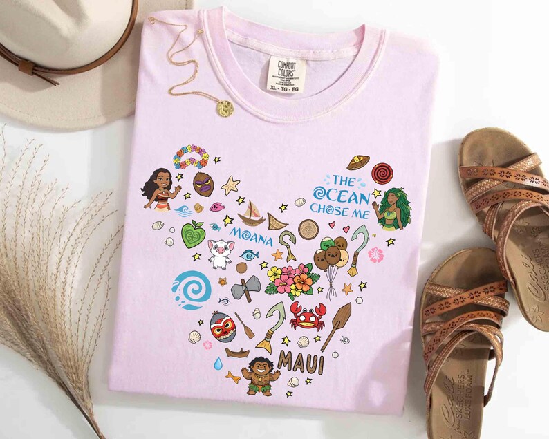 May include: Pale pink t-shirt with a heart-shaped design featuring cartoon characters and the text "MOANA" and "MAUI". The shirt also has the text "THE OCEAN CHOSE ME". A gold necklace and brown sandals are next to the shirt.