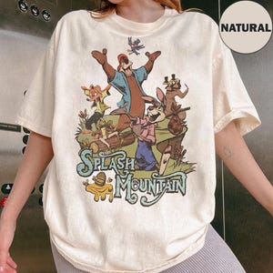 Puede incluir: Una camiseta de manga corta de color natural con un estampado gráfico de personajes de dibujos animados de Splash Mountain. El diseño incluye un oso, un conejo y un zorro en un entorno forestal con el texto Splash Mountain en una fuente estilizada.