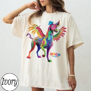 May include: An ivory short-sleeve t-shirt featuring a vibrant, multi-colored graphic of a winged alebrije dog with a long tongue. The design includes intricate patterns in shades of pink, blue, green, and yellow, with the Disney Coco logo printed below.