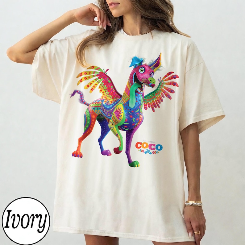May include: An ivory short-sleeve t-shirt featuring a vibrant, multi-colored graphic of a winged alebrije dog with a long tongue. The design includes intricate patterns in shades of pink, blue, green, and yellow, with the Disney Coco logo printed below.