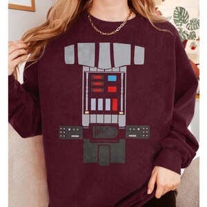 Op de afbeelding: Een bordeauxrood sweatshirt met ronde hals en een grafische print van een robotborstplaat. Het ontwerp bevat een centraal paneel met rechthoekige rode, blauwe en witte accenten, geflankeerd door twee kleinere bedieningspanelen met groene en witte details.