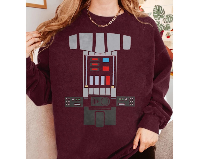Op de afbeelding: Een bordeauxrood sweatshirt met ronde hals en een grafische print van een robotborstplaat. Het ontwerp bevat een centraal paneel met rechthoekige rode, blauwe en witte accenten, geflankeerd door twee kleinere bedieningspanelen met groene en witte details.