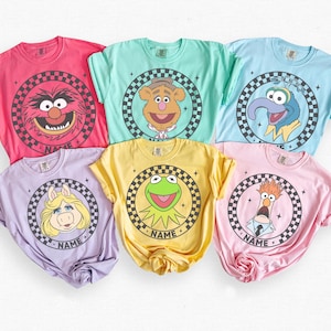 May include: Six colorful t-shirts featuring Muppet characters. Each shirt has a different character in a circular design with a checkered border and the word "NAME". Colors include pink, teal, blue, purple, yellow, and light pink.