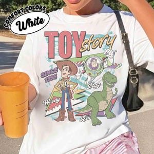 May include: White t-shirt featuring a graphic of characters from the movie "Toy Story." The design includes Woody, Buzz Lightyear, and Rex, with the words "Toy Story" and "Since 1995."
