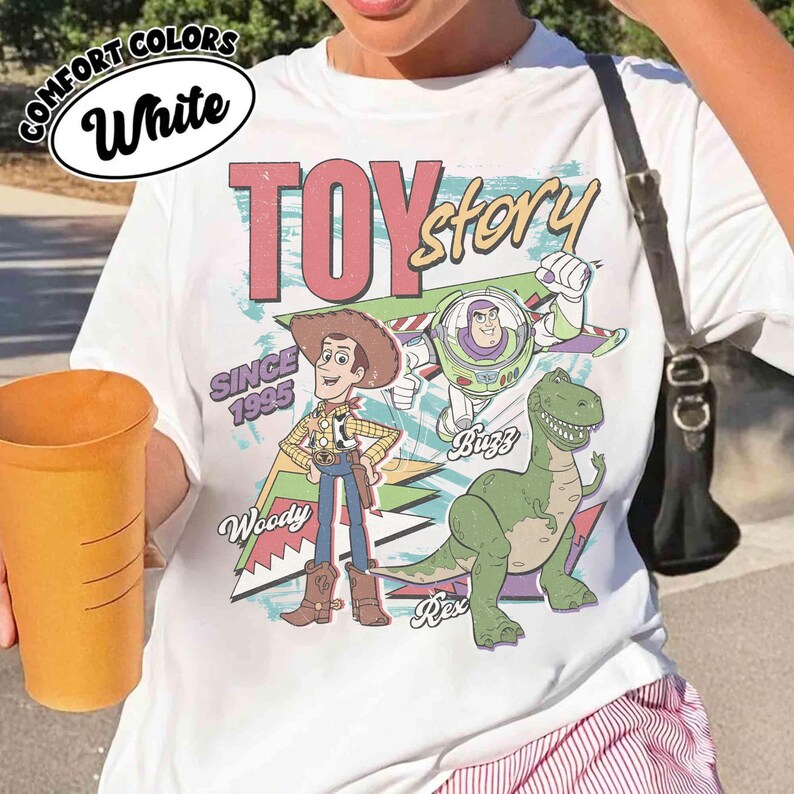 May include: White t-shirt featuring a graphic of characters from the movie "Toy Story." The design includes Woody, Buzz Lightyear, and Rex, with the words "Toy Story" and "Since 1995."