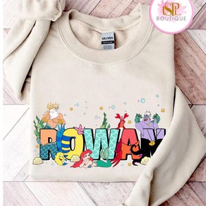 Può includere: Una felpa girocollo beige con il nome "ROWAN" in lettere colorate in stile cartone animato. Il design include illustrazioni di personaggi di un popolare film d'animazione, con il logo del marchio in alto a destra.