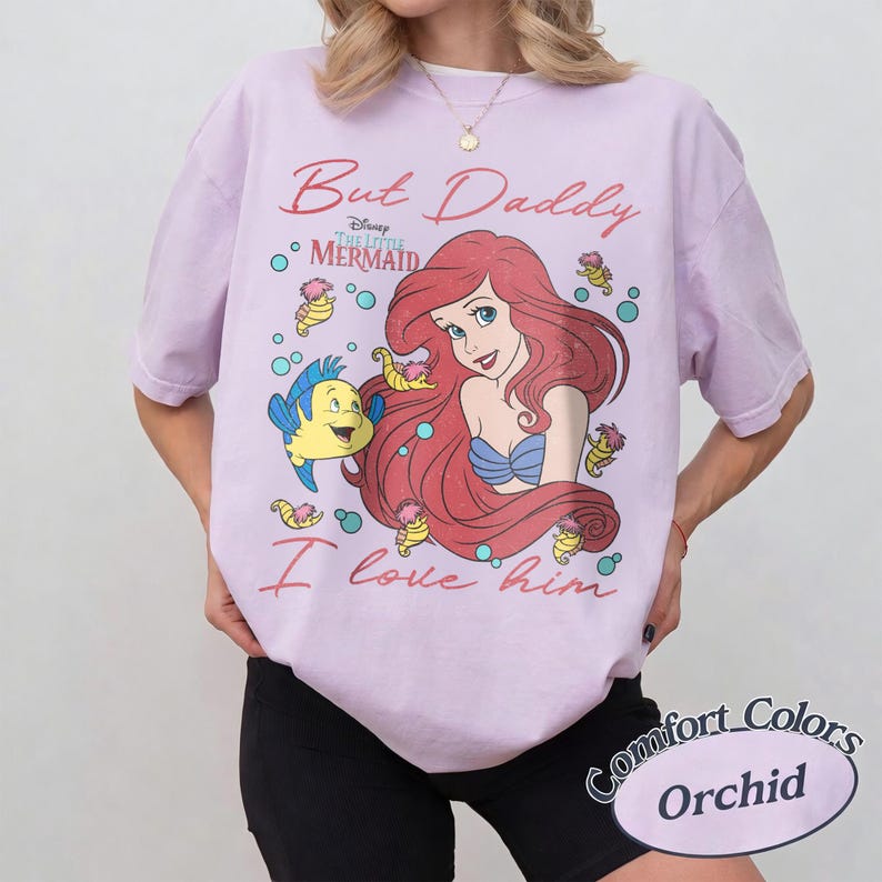 May include: A light purple t-shirt featuring a graphic of Ariel and Flounder from The Little Mermaid. The text reads But Daddy I love him. The design includes small seahorses and bubbles surrounding the characters. The shirt has a crew neckline.