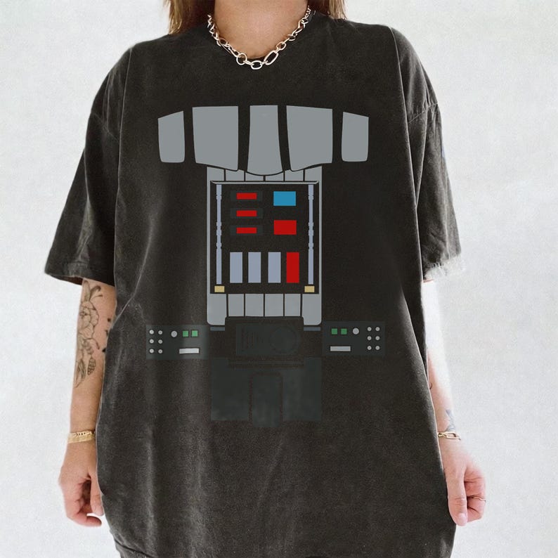 Op de afbeelding: Een antracietgrijs T-shirt met korte mouwen en een grafische print van een bedieningspaneel op de borst met grijze, rode en blauwe rechthoekige accenten. Het ontwerp bootst een mechanische pakinterface na met een centrale console.