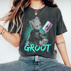May include: Dark gray t-shirt featuring a graphic of Groot holding a pink cassette tape. The word "GROOT" is written in large, turquoise letters below the character. The design is set against a turquoise dotted background.