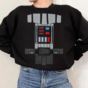 Op de afbeelding: Een zwart sweatshirt met lange mouwen en een grafische print op de borst die een mechanisch bedieningspaneel nabootst. Het ontwerp bevat grijze rechthoekige vormen, een centraal raster met rode, blauwe en witte vierkanten en zijpanelen.
