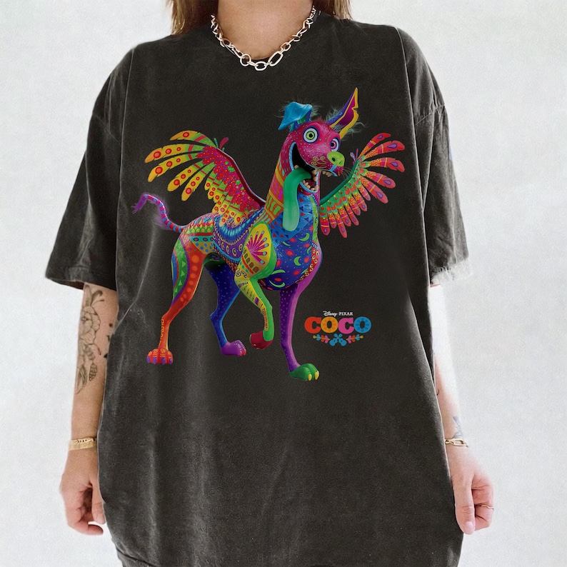 May include: A charcoal gray short-sleeve t-shirt featuring a vibrant, multi-colored graphic of Dante the alebrije dog from the movie Coco. The dog has wings, a long tongue, and intricate patterns in shades of pink, green, blue, and yellow.