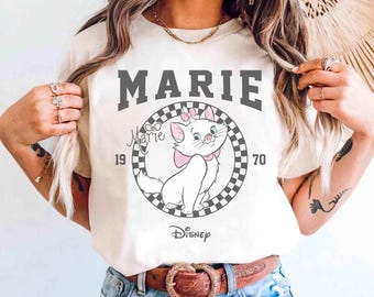 May include: Cream-colored t-shirt featuring a graphic of Marie, the white kitten from Disney's Aristocats. The design includes the name "MARIE" in gray block letters, the year "1970", and the Disney logo. The kitten has a pink bow.