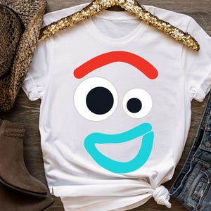 May include: White t-shirt with a cartoon face design. It has a red eyebrow, two eyes, and a blue smile. The shirt is displayed with a cowboy hat, boots, and denim shorts. The design is simple and colourful.