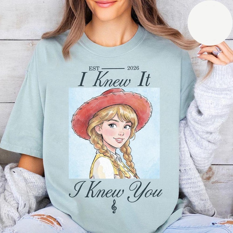 May include: A light blue t-shirt featuring a graphic illustration of a blonde character with braided hair wearing a red cowboy hat. The text reads EST 2026, I Knew It, and I Knew You, with a small musical treble clef symbol at the bottom.