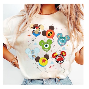 May include: Cream-coloured t-shirt featuring cartoon character balloons. The balloons are designed as Mickey Mouse heads, with characters like Woody, Mike Wazowski, and Lightning McQueen. The design includes stars and the number 95.