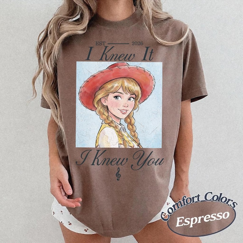 May include: A brown short-sleeve t-shirt featuring a graphic of a blonde character wearing a red cowboy hat and braids. The text reads EST 2026 I Knew It I Knew You with a small musical note symbol. The design is centered on the chest area.
