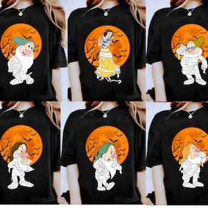 以下が含まれることがあります： ハロウィーンをテーマにした漫画のキャラクターが描かれた黒いTシャツ。各シャツには、コウモリのいるオレンジ色の月の背景に、ミイラの衣装を着たさまざまなキャラクターが表示されます。キャラクターには、白雪姫と7人の小人が含まれます。