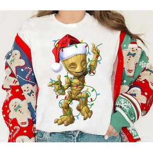 May include: White t-shirt featuring a cartoon Groot character wearing a Santa hat and wrapped in Christmas lights. The sleeves of the shirt are visible, with a red and blue gingerbread man pattern on one side and a green and red pattern on the other.