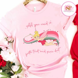 May include: Pale pink t-shirt with a cartoon fairy design and the words "All you need is faith trust and pixie dust." The fairy is depicted with a wand and a rainbow. The shirt has a round neck and short sleeves. The design uses pink, green, and yellow.
