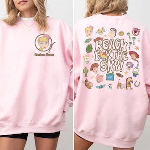 May include: A light pink crewneck sweatshirt featuring a small graphic of a blonde character in a lasso on the front left chest with the text Custom Name. The back displays various cartoon characters and the text REACH FOR THE SKY!