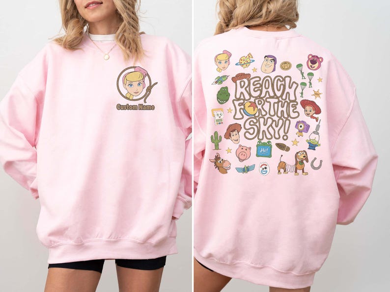 May include: A light pink crewneck sweatshirt featuring a small graphic of a blonde character in a lasso on the front left chest with the text Custom Name. The back displays various cartoon characters and the text REACH FOR THE SKY!
