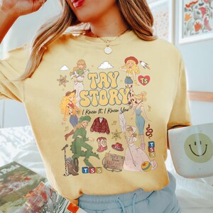 May include: A pale yellow crewneck t-shirt featuring a graphic print with the text TAY STORY I Knew It, I Knew You. The design includes various illustrated figures, a red cardigan, cowboy boots, a hat, blocks labelled TS 5, and a sign reading Tay's Room Keep Out.