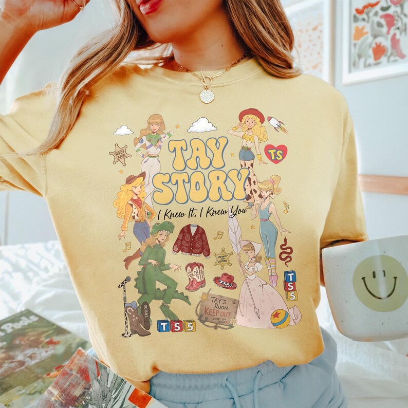 May include: A pale yellow crewneck t-shirt featuring a graphic print with the text TAY STORY I Knew It, I Knew You. The design includes various illustrated figures, a red cardigan, cowboy boots, a hat, blocks labelled TS 5, and a sign reading Tay's Room Keep Out.