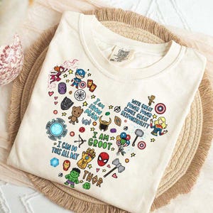 Pode incluir: Uma t-shirt de manga curta cor de creme com uma colagem em forma de coração de vários ícones, personagens e citações de super-heróis, incluindo "I am Iron Man", "I am Groot", "I can do this all day" e "With great power comes great responsibility".