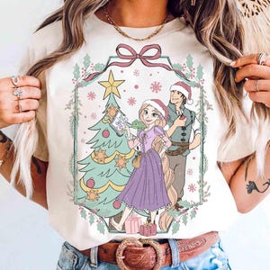 May include: Cream-colored t-shirt featuring a Christmas-themed illustration. The design includes a Christmas tree, a couple wearing Santa hats, and gift boxes. The artwork is framed with a pink bow and holly.