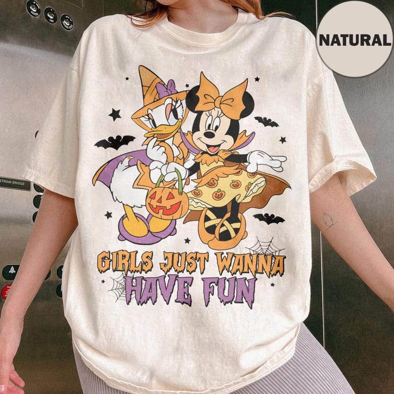 May include: A natural-colored t-shirt featuring a graphic of Daisy Duck and Minnie Mouse dressed in Halloween costumes. The text reads GIRLS JUST WANNA HAVE FUN. The design includes pumpkins, bats, and spiderwebs in orange, purple, and black colors.