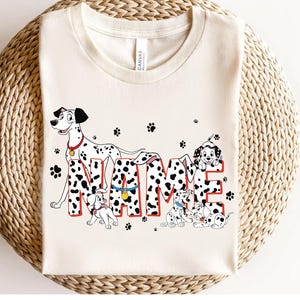 Puede incluir: Camiseta color crema con un diseño de perro dálmata. La palabra "NAME" está escrita con un patrón dálmata y contornos rojos. También se incluyen varios cachorros dálmatas y huellas de patas.